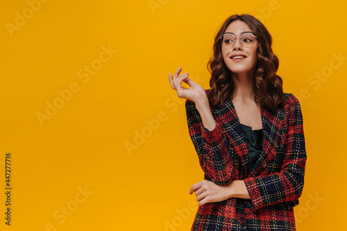 Charming woman with curly hair in eyeglasses posing on isolated backdrop. Brunette girl in red striped jacket smiling on yellow background..