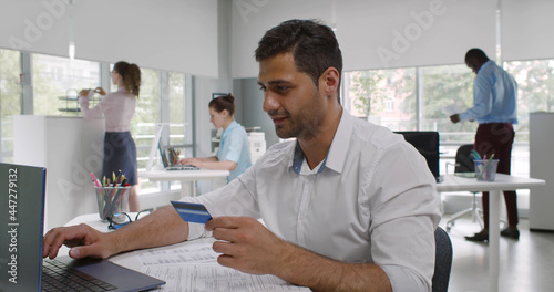 Indian businessman sitting at desk in office holding credit card and paying online on laptop