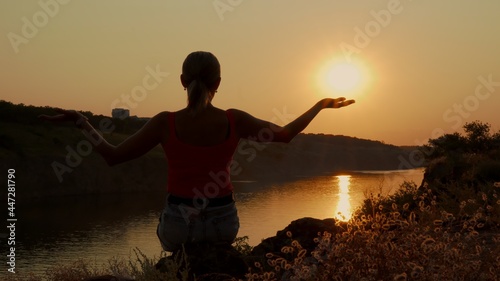The silhouette of a woman sitting on the top of a mountain with her arms raised, in one of which she seems to hold the sun. The exposure of the shot enhances the beauty of the sunset.