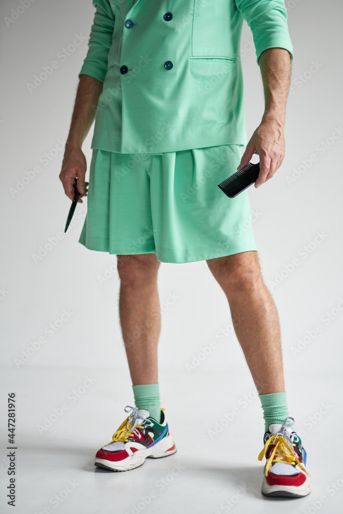 Cropped shot of trendy middle aged man wearing colorful stylish outfit, holding sharp barber scissors and hair comb while standing over white background