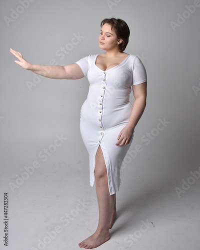 Full length portrait of young plus sized woman with short brunette hair,  wearing a tight white body con dress,   standing walking  posewith light studio background. photo