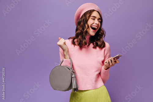 Happy lady in pink beret, sweater and green skirt smiles and reads messages on phone. Cheerful gl holds grey handbag on purple background.