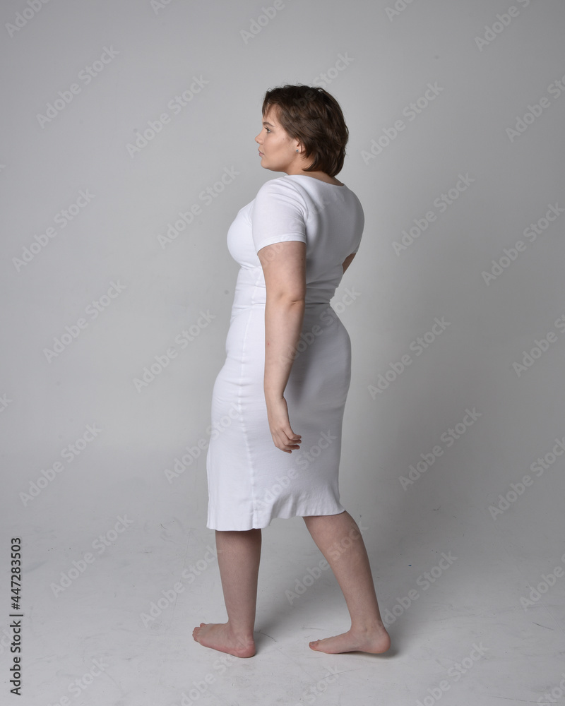 Full length portrait of young plus sized woman with short brunette hair,  wearing a tight white body con dress,   standing walking  posewith light studio background.