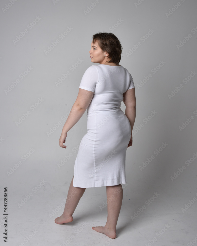 Full length portrait of young plus sized woman with short brunette hair, wearing a tight white body con dress, standing walking posewith light studio background. Full length portrait of young plus sized woman with short brunette hair, wearing a tight white body con dress, standing walking posewith light studio background.