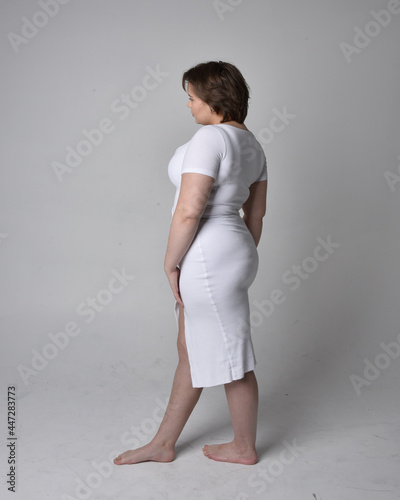 Full length portrait of young plus sized woman with short brunette hair,  wearing a tight white body con dress,   standing walking  posewith light studio background. photo