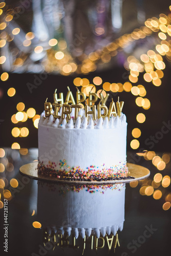Birthday cake with candles, garland with bright bokeh lights on the background. The white cake is decorated with colored sprinkles and stands on a reflective surface.