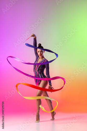 Beautiful young female rhythmic gymnast posing with bright ribbon in her hand isolated on multicolored neon background. photo