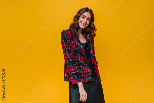 Curly woman in red jacket smiling on yellow background. Happy girl with brunette hair in striped clothes looking into camera on isolated backdrop..