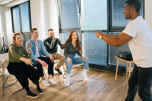 Group of young diverse multiethnic colleagues playing in active games during team building in modern office room. Cheerful African American male playing charades with friends showing pantomime.