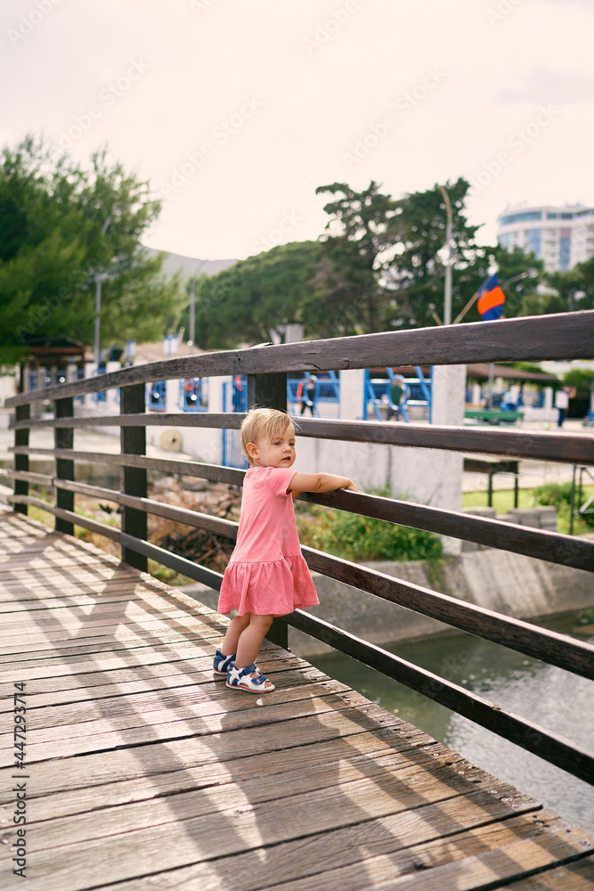 Little girl stands on the bridge above the water, leaning on the handrail with her hands