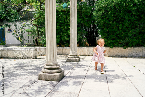 Little girl walks along a tiled path near the columns against the background of green magnolia bushes © Nadtochiy