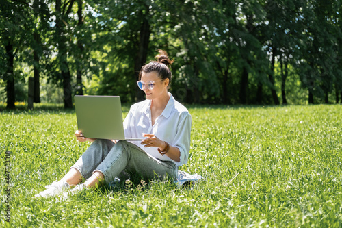 attractive business women in glasses and white blouse using laptop sitting on grass on break.