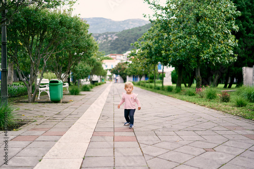 Little girl walking along the paved road in the park against the background of green trees and mountains