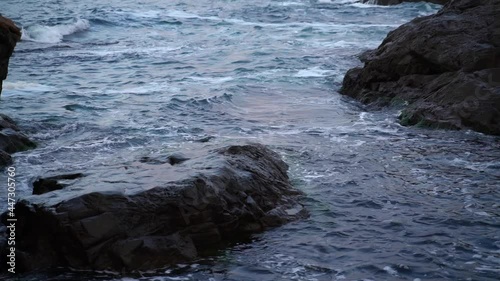 Gentle blue sea waves rushing and splashing against dark gray cliffs forming white foam. Slow motion