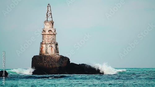 Sea waves hitting and splashing against old white lighthouse and dark gray cliffs. Isolated on blue cloudy sky. Slow motion. Ahtopol on Black sea coast, Bulgaria