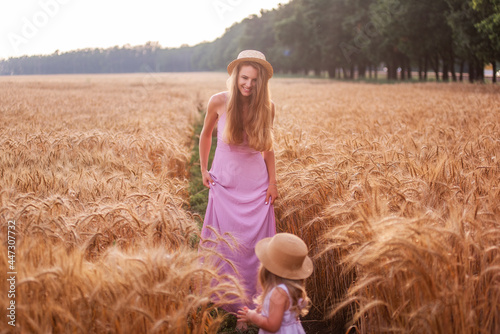 Daughter hold mother s hand  walk along the golden wheat field at sunset. Young woman and a girl in pink dresses and straw hats stand among the rye  their feet in the mud. Agro walk in the countryside