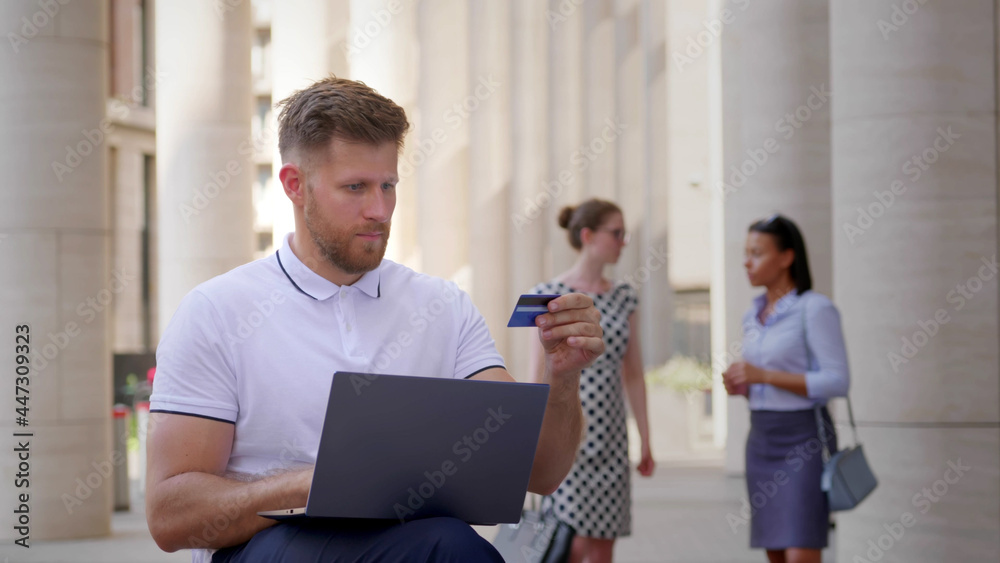 Medium shot of caucasian businessman using laptop and credit card outdoors office building Medium shot of caucasian businessman using laptop and credit card outdoors office building