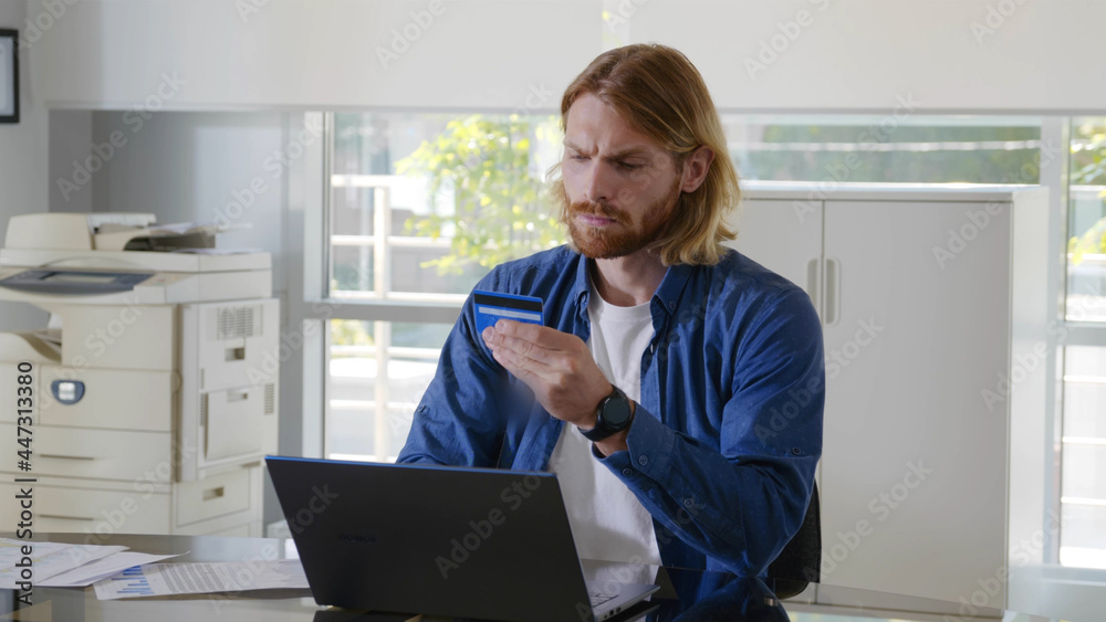 Young confident businessman sitting at desk paying on computer with credit card in office Young confident businessman sitting at desk paying on computer with credit card in office