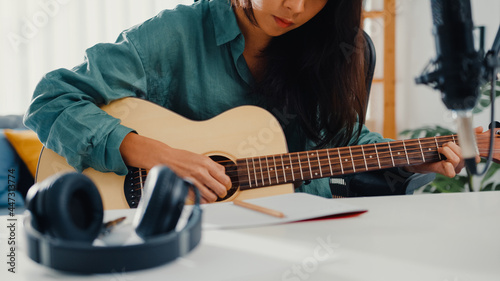 Happy asia woman songwriter play acoustic guitar listen song from smartphone think and write notes lyrics song in paper sit in living room at home studio. Music production at home concept.