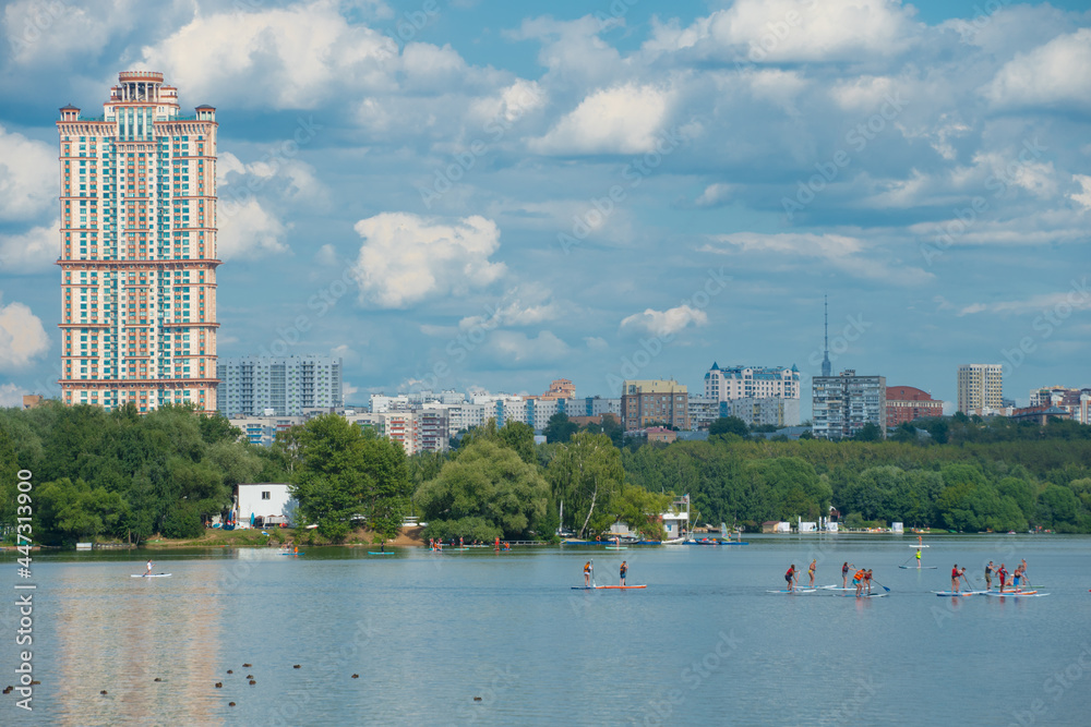 Group of SUP boards in the distance on the Stroginsky Bay of Moskva river float against the backdrop of city buildings and a skyscraper, summer weekend in Moscow
