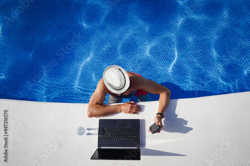 bird view of remote online working digital nomad man on workation with hat & laptop at a white table standing in a sunny turquoise water pool