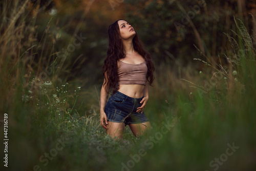 Sexy woman posing in a field in the grass rays of the sunset. Lifestyle on nature dressed in jeans shorts and beige top in golden hour. Romantic autumn mood, long tall grass in the meadow © oleg_ermak