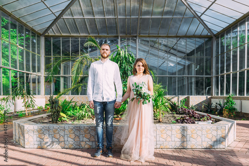 The bride and groom walk in the botanical garden near the ancient glass architecture, hold on to hands