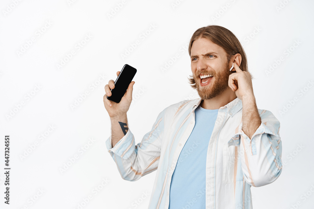 Image of happy blond guy dancing with earphones, listening music in headphones, holding mobile phone and smiling pleased, standing over white background Image of happy blond guy dancing with earphones, listening music in headphones, holding mobile phone and smiling pleased, standing over white background