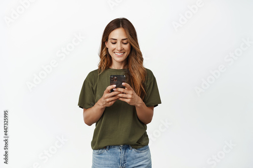 Happy smiling woman, adult female looking at her mobile phone, chat in application, sending message, using online shopping app, standing against white background