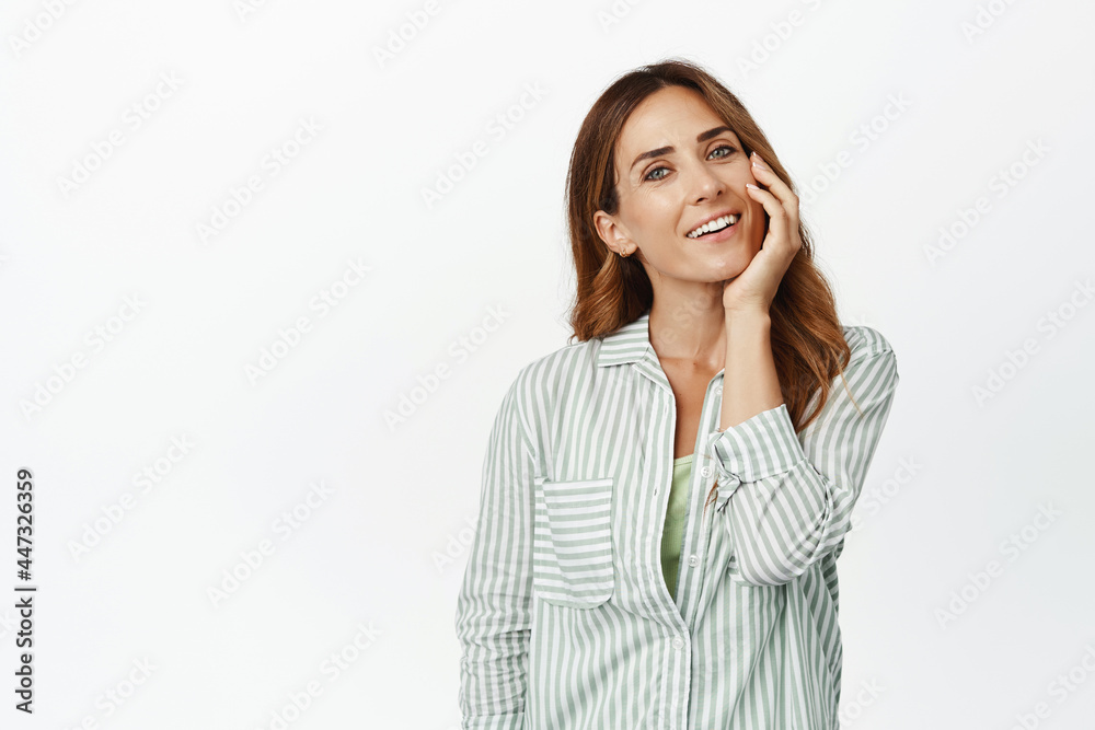 Middle aged woman touching her face with pleased smile, enjoying softness of clean facial skin after skincare cosmetics, standing against white background Middle aged woman touching her face with pleased smile, enjoying softness of clean facial skin after skincare cosmetics, standing against white background