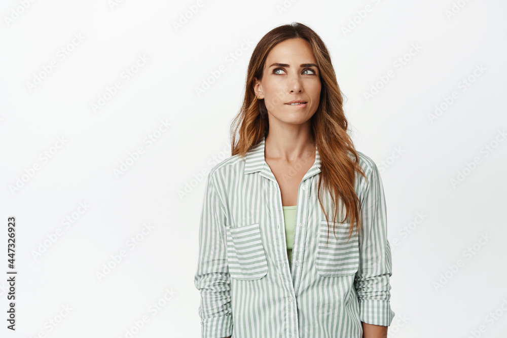 Image of thoughtful woman squinting, biting lip and thinking, looking away while scheming, imaging smth interesting, has idea, standing against white background Image of thoughtful woman squinting, biting lip and thinking, looking away while scheming, imaging smth interesting, has idea, standing against white background