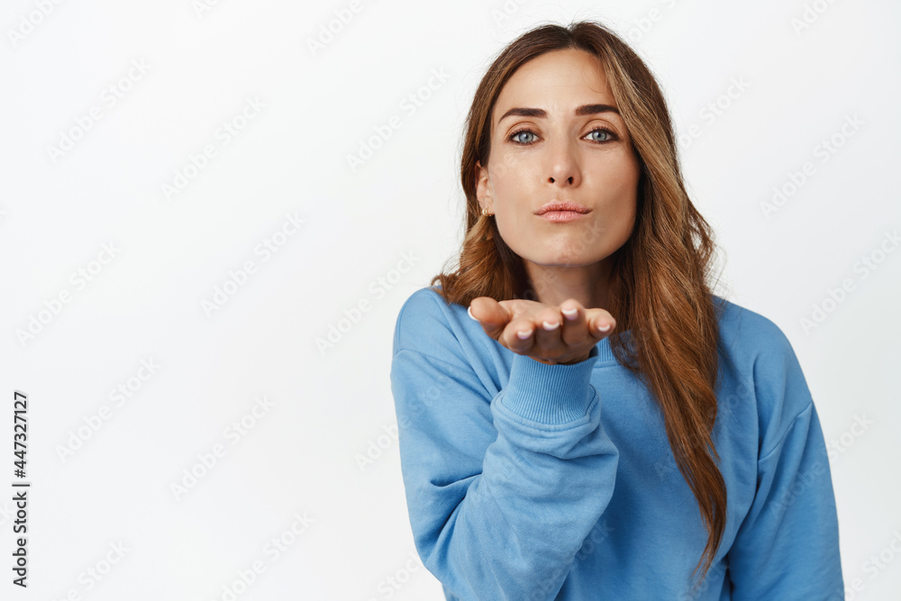 Tender and feminine woman blowing air kiss, holing open hand near puckered lips and gazing cute at camera, standing against white background Tender and feminine woman blowing air kiss, holing open hand near puckered lips and gazing cute at camera, standing against white background