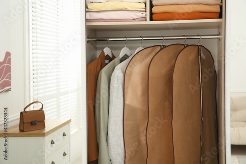 Photo Garment bags with clothes on rack in wardrobe indoors