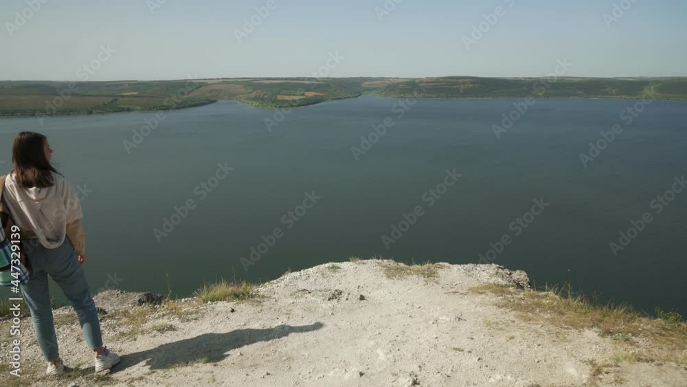Attractive female hiker with backpack walking on edge of high rock near Dniester river. Beautiful view of Bakota bay on background. Active lifestyle concept. Attractive female hiker with backpack walking on edge of high rock near Dniester river. Beautiful view of Bakota bay on background. Active lifestyle concept.