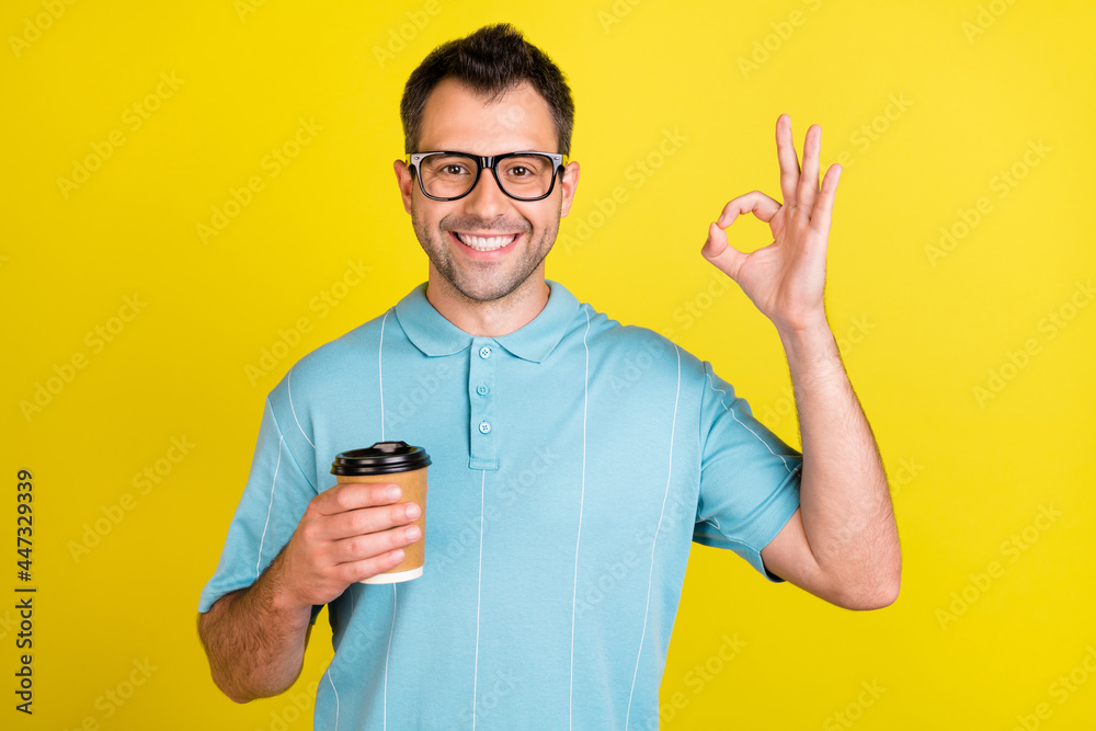 Photo of cool brunet man hold coffee show okey sign wear eyewear blue t-shirt isolated on yellow color background Photo of cool brunet man hold coffee show okey sign wear eyewear blue t-shirt isolated on yellow color background