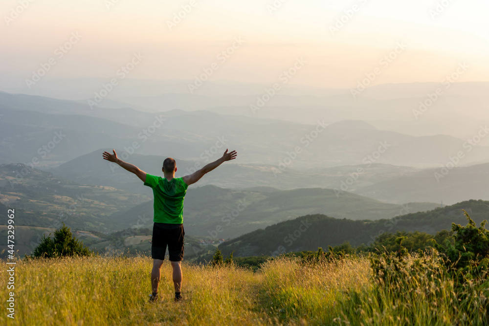 Fototapeta premium Happy man standing alone against the backdrop of a mountain landscape with his hands raised high. Summer, sun, dawn.