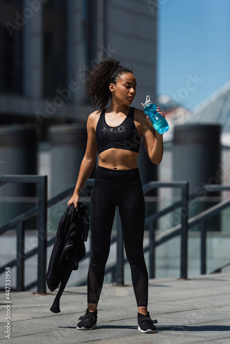 African american sportswoman holding jacket and water in sports bottle outdoors