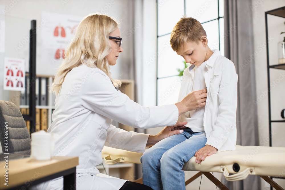 Fototapeta premium Qualified female doctor in lab coat and eyeglases using stethoscope fo listening boy's lungs during medical checkup. Control of health condition. Medicine concept.