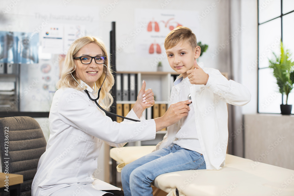 Healthcare and pediatrics concept. Pretty mature smiling professional woman physician checking teen boy's lungs and heart with stethoscope, gesturing together thumbs up into camera