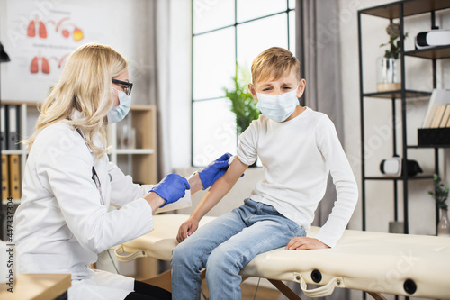 Healthcare and vaccination concept. Professional female doctor in medical mask doing vaccine injection in teen boy's arm. Boy with scared eyes, in protective mask sitting on the couch