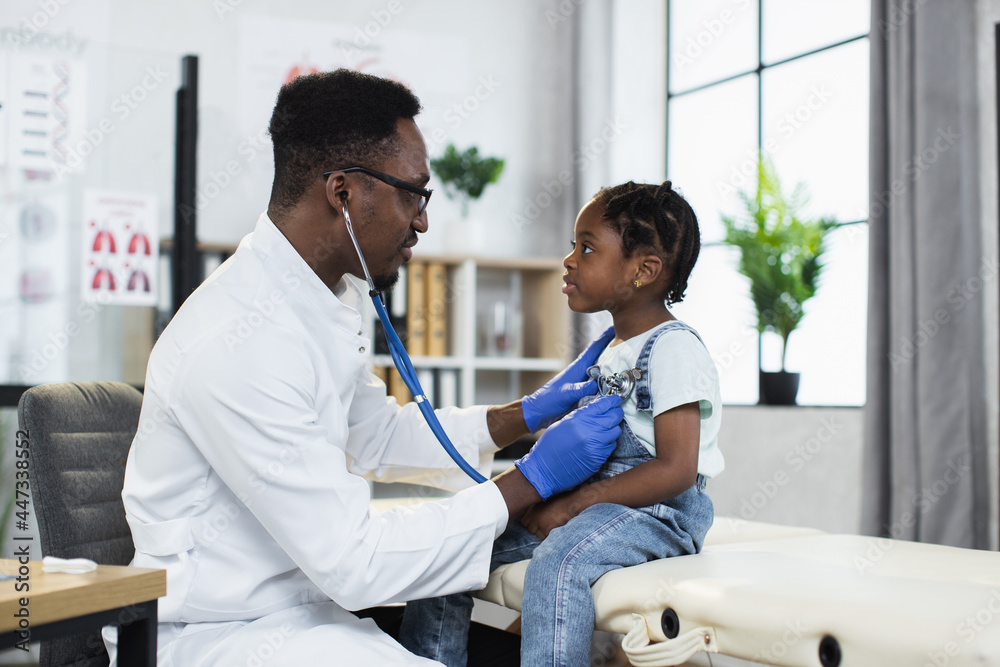 Side view of african american pediatrician using stethoscope for listening little girl's lungs at modern hospital. Qualified doctor doing checkup for female patient. Side view of african american pediatrician using stethoscope for listening little girl's lungs at modern hospital. Qualified doctor doing checkup for female patient.