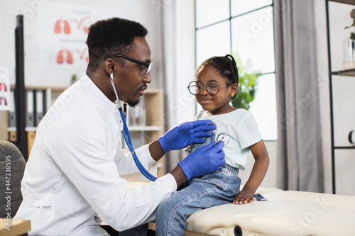 Pediatrics and regular check up concept. Pleasant caring Afro-American man physician checking littel African kid girl s heartbeat and breath using stethoscope  working in medical center
