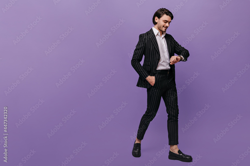 Attractive young man in white shirt, black striped suit and trendy shoes smiling and looking at watch on hand. Lifestyle and business concept against violet background Attractive young man in white shirt, black striped suit and trendy shoes smiling and looking at watch on hand. Lifestyle and business concept against violet background
