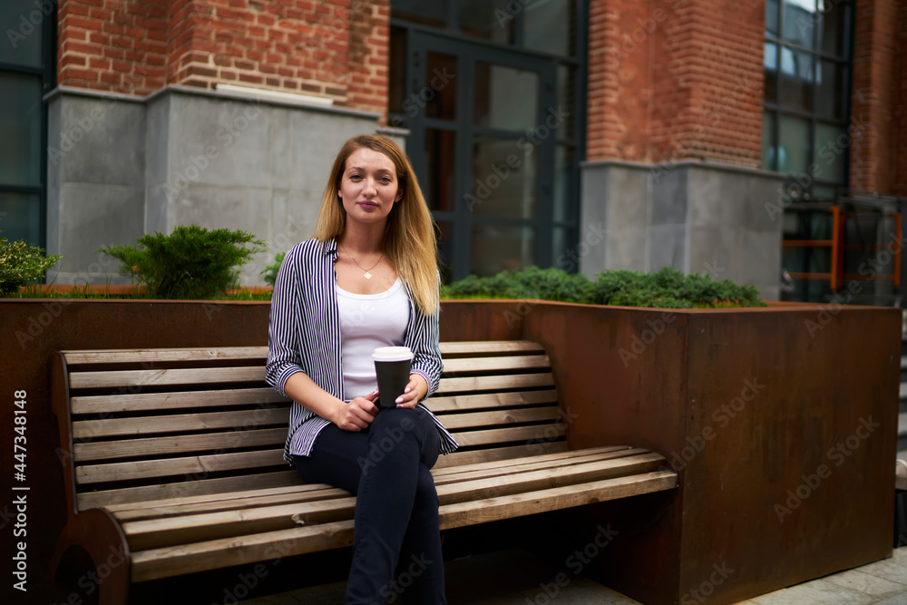 Obraz premium Portrait of attractive female student with disposable takeaway cup resting at city bench and looking at camera during coffee break in campus, beautiful hipster girl with caffeine beverage posing