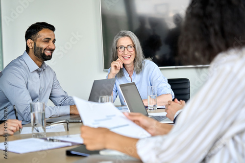 Senior female ceo and happy multicultural business people discussing company presentation at boardroom table. Smiling diverse corporate team working together in modern meeting room office.