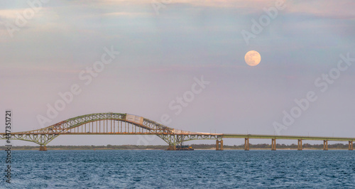 Full moon rising through a soft pastel colored sky above a long steel tied arch bridge. Fire Island Inlet Bridge, New York