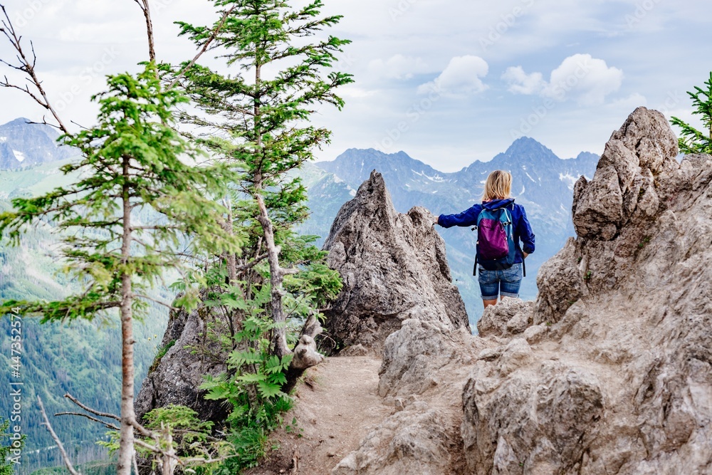 Fototapeta premium Back view of a blond woman traveler standing between rocks in the mountains. She admires the beautiful mountain views.
