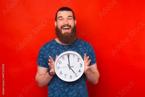 A nice photo of a young man holding a white clock is smiling at the camera near a red wall