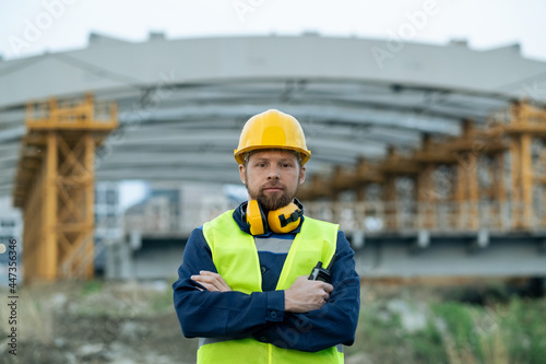 Portrait of foreman in headphones and in work helmet standing with arms crossed on construction site and looking at camera