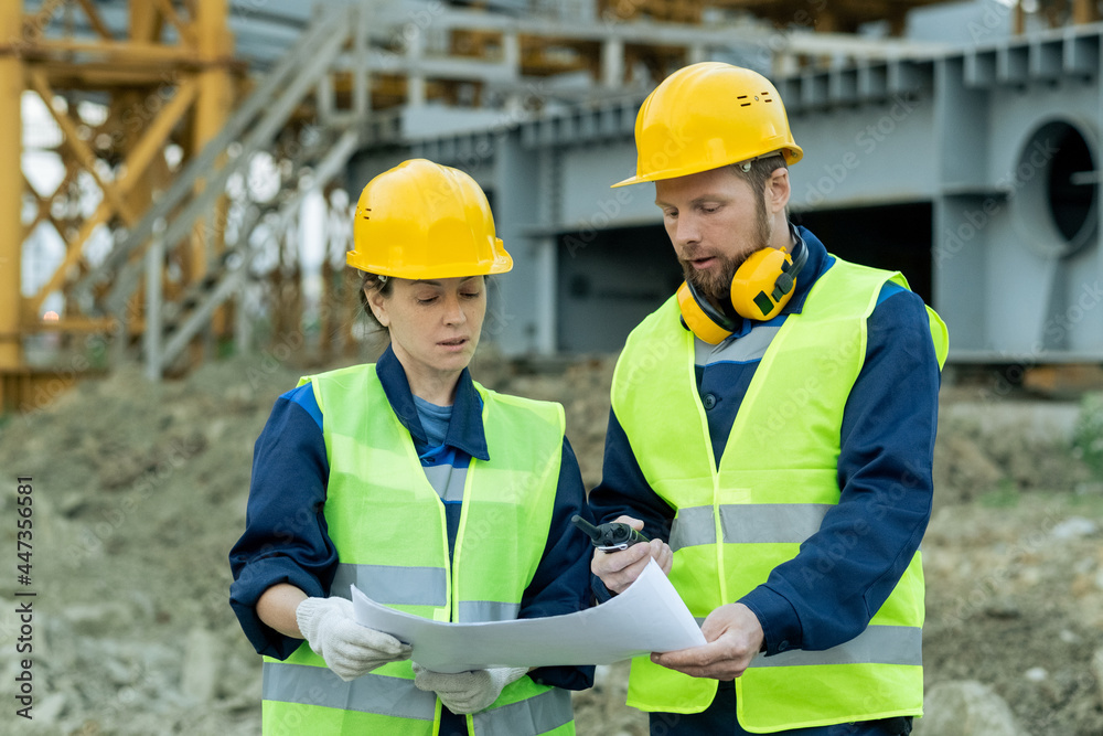 Two engineers in workwear and in work helmets examining blueprint while working in team on construction site Two engineers in workwear and in work helmets examining blueprint while working in team on construction site
